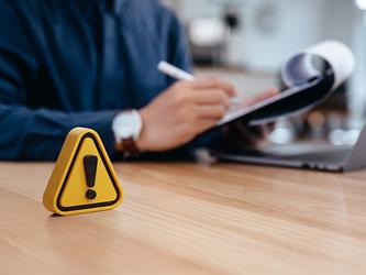 yellow triangle with exclamation mark on a desk, with person with clipboard in background